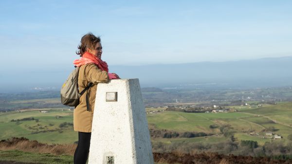 A woman with a coral scarf stands at a trig point looking across open countryside.